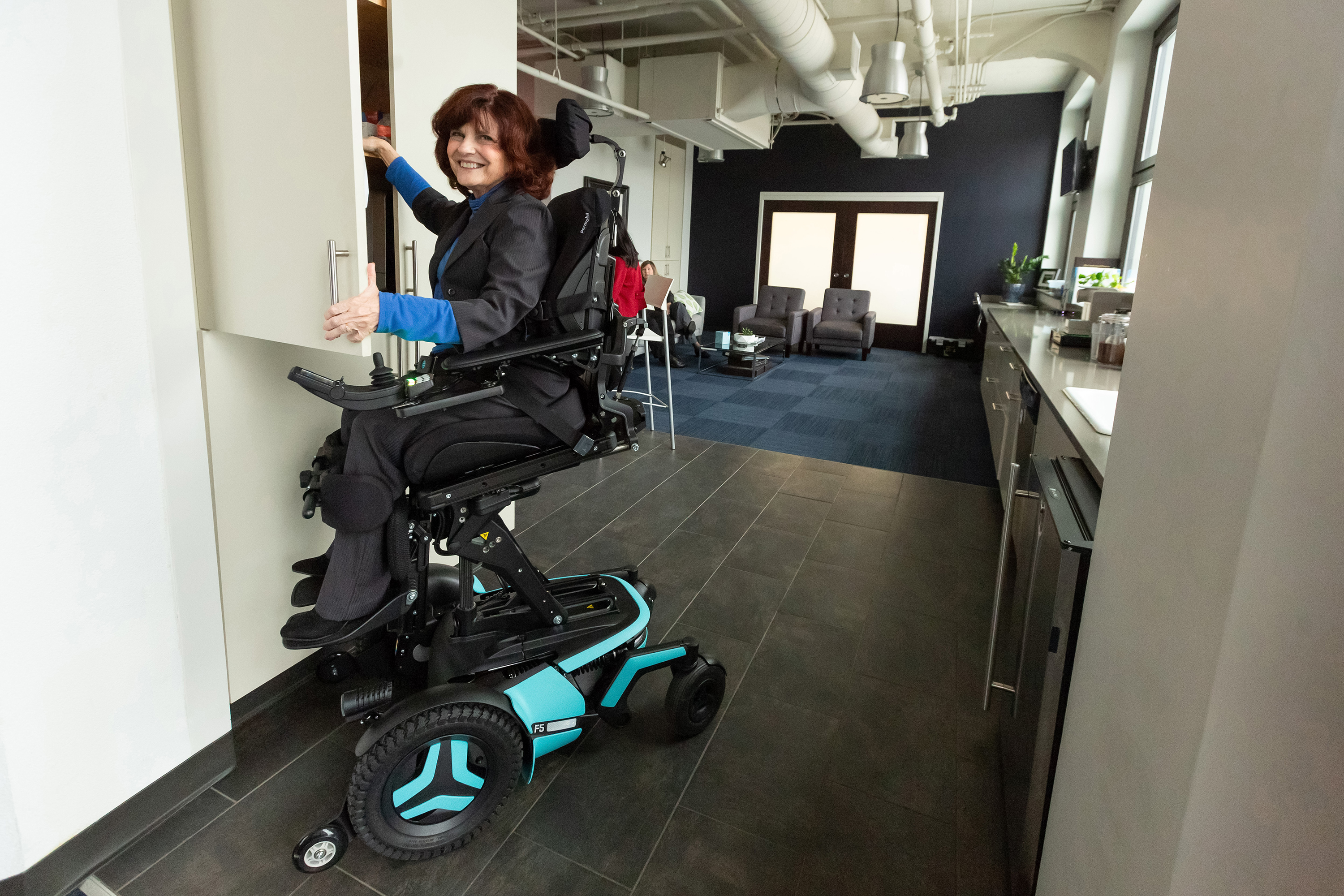 A Caucasian woman smiles and faces the camera. She is using her F5 power chair with light blue accents in ActiveReach position to access her upper cabinets.