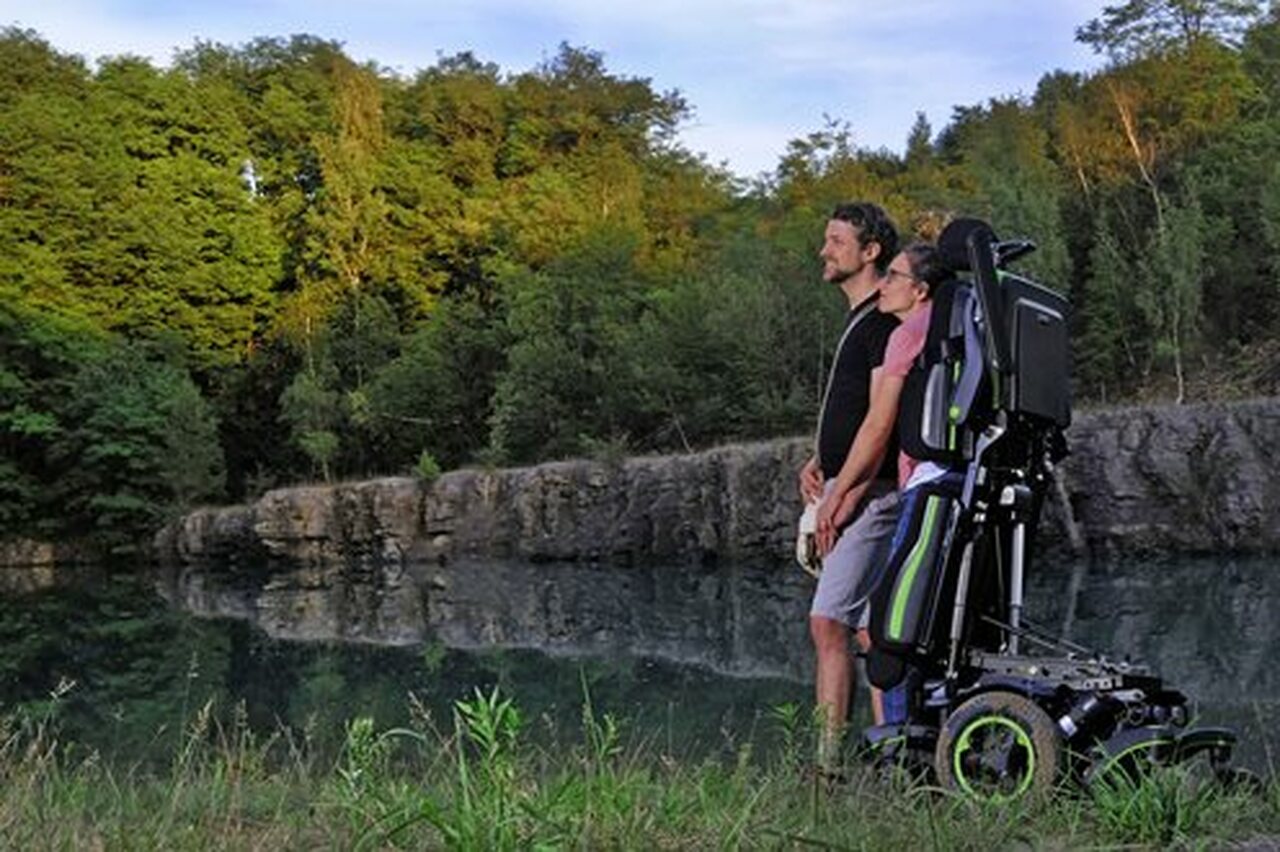 A woman using the Q700-UP standing wheelchair stands close to a man by a lake with green trees in the background