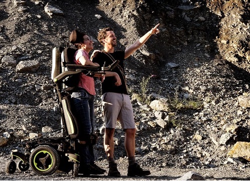 A woman in a pink tank top, using the Q700-UP standing wheelchair stands close beside a man. They are in front of a rocky cliff and the man is smiling and pointing at something in the distance.