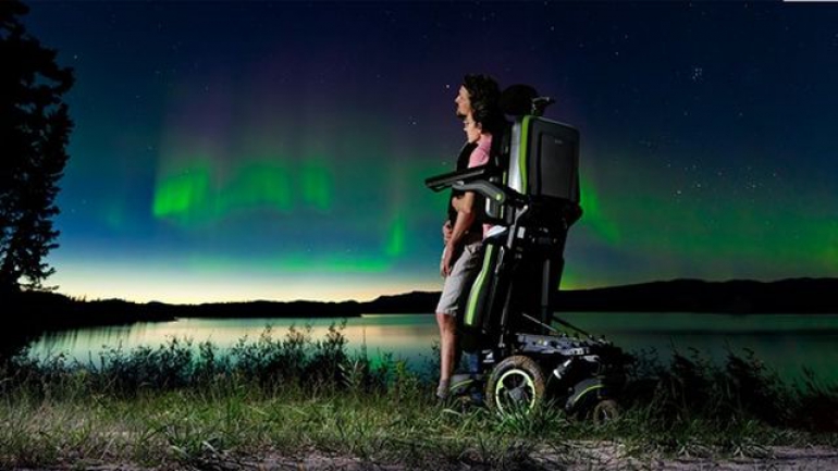 A woman using the Q700-UP standing wheelchair stands close to a man in a field near a lake. It's nighttime and they are watching the Northern Lights.