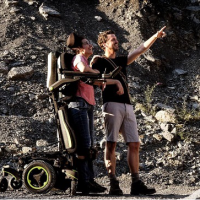 A woman in a pink tank top, using the Q700-UP standing wheelchair stands close beside a man. They are in front of a rocky cliff and the man is smiling and pointing at something in the distance. thumbnail