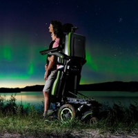 A woman using the Q700-UP standing wheelchair stands close to a man in a field near a lake. It's nighttime and they are watching the Northern Lights. thumbnail