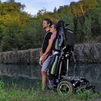 A woman using the Q700-UP standing wheelchair stands close to a man by a lake with green trees in the background thumbnail