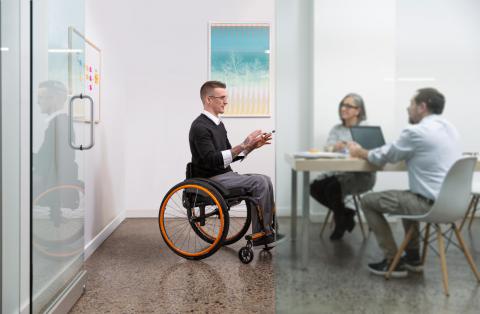 A person in an Apex wheelchair with orange detailing meets with two others at a table. They are gesturing with their hands and the other two people are listing intently.