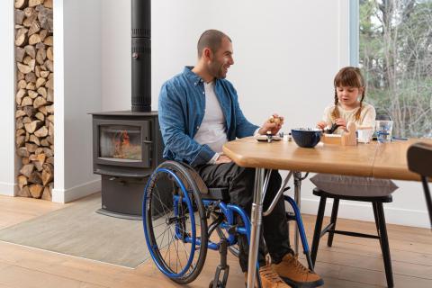A man using a Helio C2 wheelchair sits at a table with his daughter, eating breakfast. There is a wood burning stove and a stack of chopped firewood in the background.